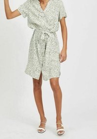 Femme portant une robe courte à manches courtes verte claire avec des taches abstraites blanches, cintrée à la taille, et des sandales blanches à bout ouvert.