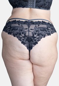 Schwarze Spitzenunterhose mit floralen Mustern, durchscheinendem Material, gewellten Rändern und dünnen Trägern auf der Rückseite. Die strukturierte Oberfläche hebt das Design hervor.