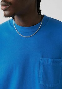 Blue short-sleeve t-shirt with a textured fabric, round neckline, and a left chest pocket featuring embossed branding. Silver chain necklace visible.