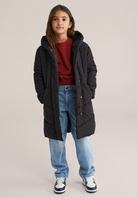 Jauns cilvēks stāv, ģērbies melnā garā pūkā jakā, sarkanā džemperī, brīvās zilo džinsu biksēs un melnbaltās sporta kurpēs uz vienveidīga fona.