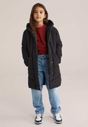 Joven de pie, vistiendo una chaqueta de plumas larga y negra, un suéter rojo, jeans azul holgados y zapatillas negras y blancas sobre un fondo liso.