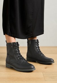 Bottes en cuir noir mi-mollet à lacets avec fermetures éclair latérales portées avec une jupe noire sur un sol en bois.