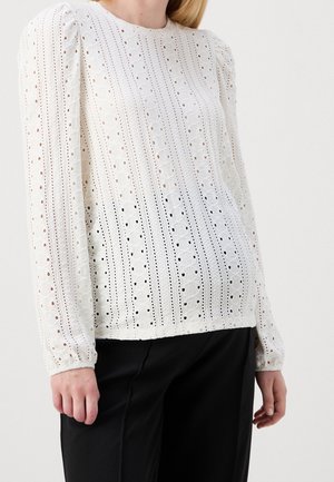 Weiße Langarmbluse mit vertikalen Lochspitzenmustern, getragen zu schwarzen Hosen, zeigt eine lockere Passform und einen gerundeten Ausschnitt.