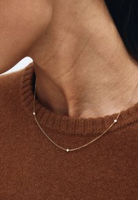 Zlatý retiazkový náhrdelník s jemnými očkami a malými bielymi korálkami, nosený cez hnedý rebrovaný sveter. Jednoduchý dizajn s jemnými detailmi.