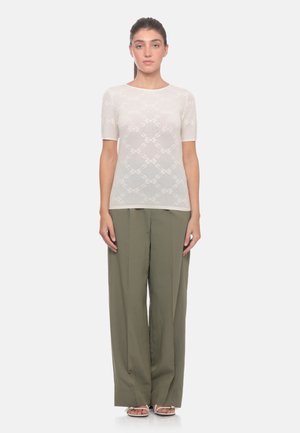 Donna in piedi che indossa una maglietta bianca trasparente a maniche corte con motivo a diamante e pantaloni larghi verde oliva con sandali.