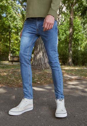 Personne portant un jean skinny bleu clair et des baskets montantes blanches, debout sur un chemin pavé dans un parc avec des arbres et un banc à proximité.