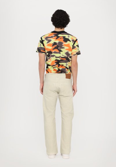 Short-sleeve t-shirt with a multicolor camouflage pattern featuring orange, green, and black. Paired with light beige straight-leg pants.