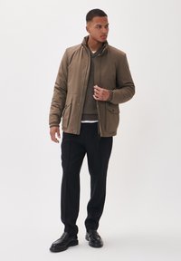 Giacca imbottita marrone con chiusura a zip, due tasche frontali, colletto alto, indossata sopra un maglione marrone e una camicia bianca, abbinata a pantaloni neri.