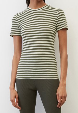 Nainen, jolla on lyhythihainen valkoinen ja oliivinvihreä vaakaraitainen t-paita ja oliivinvihreät ribbineulostrikoot, seisoo valkoisen seinän edessä.