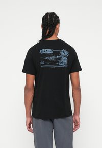 Crna pamučna majica s plavim grafičkim dizajnom na leđima, koja prikazuje palmu i pejzaž s natpisom "RIP CURL Surfing Co."