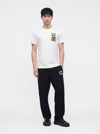 Νέος άνδρας που φοράει λευκό T-shirt Moschino με γραφικό αρκούδας και μαύρο παντελόνι φόρμας Moschino, στέκεται μπροστά σε απλό λευκό φόντο.