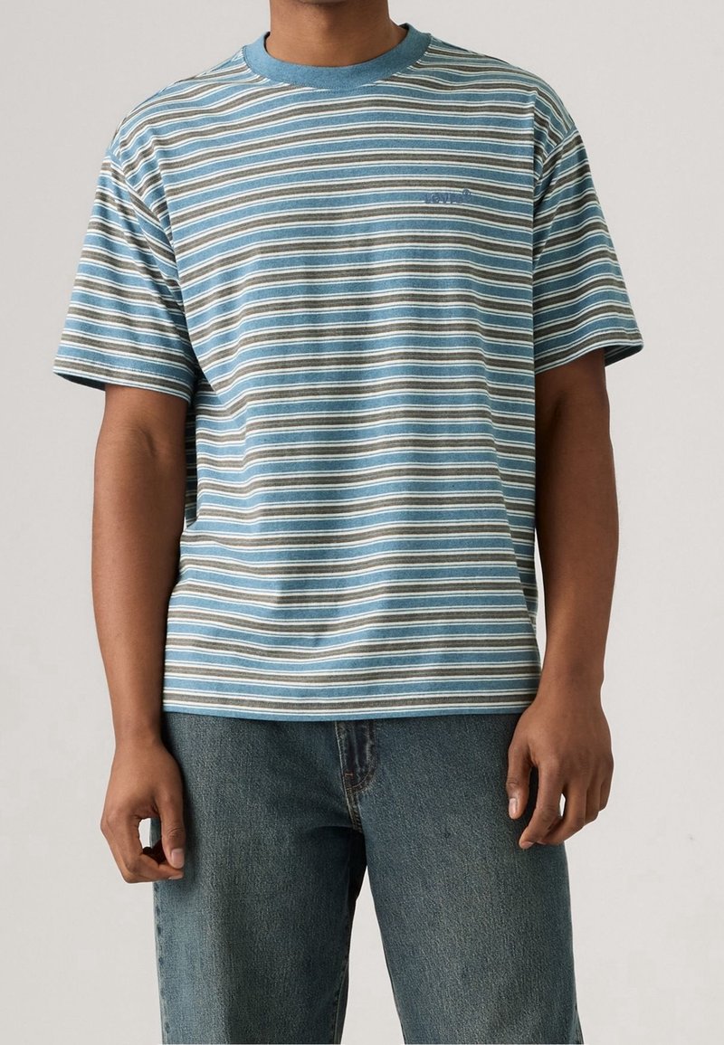 Homme portant un t-shirt à rayures bleues, blanches et marron à manches courtes avec un col bleu et un jean bleu, debout devant un fond uni.