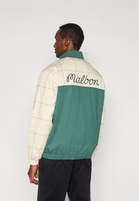 Πράσινο και μπεζ μπουφάν με καρό μοτίβο, ελαστική μέση και κεντημένο "Malbon" στην πλάτη. Κατασκευασμένο από ελαφρύ ύφασμα.