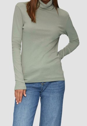 Kvinde iført lysegrøn langærmet rullekrave og blå jeans, stående med den ene hånd på hoften og den anden hvilende på låret.