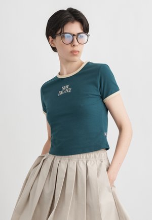 Νεαρό άτομο με κοντά σκούρα μαλλιά και στρογγυλά γυαλιά, φορώντας ένα κομψό κορυφαίο μπλουζάκι της New Balance σε τιρκουάζ χρώμα και μπεζ πλισέ φούστα, με τα χέρια στις τσέπες.