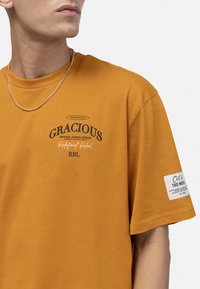 T-shirt à manches courtes moutarde ornée de texte noir imprimé, col rond, et d'une petite étiquette blanche sur la manche contenant des détails de marque supplémentaires.