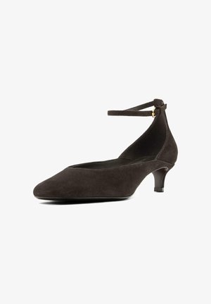 Dècolletè marrone scuro in suede con cinturino alla caviglia, punta a punta e tacco basso e spesso. Caratterizzato da una silhouette elegante e un dettaglio con fibbia laterale.