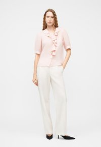 Γυναίκα με κυματιστά κόκκινα μαλλιά που φοράει ένα ανοιχτό ροζ μπλουζάκι με κοντά μανίκια και βολάν μπροστά, και άσπρο παντελόνι με φαρδιά μπατζάκια, συνδυασμένο με μαύρες γόβες.