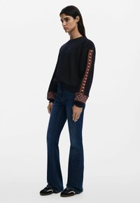 Felpa marina con ricamo geometrico rosso e bianco sulle maniche, abbinata a jeans svasati blu scuro e sneakers nere.