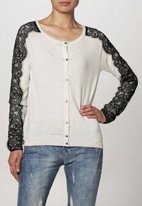 Kvinna iförd vit knapplistad kofta med svarta spetsärmar och blå slitna jeans, stående med ena handen på höften.