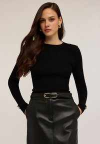 Femeie cu păr lung, brunet, purtând cercei din aur în formă de cerc, un pulover negru cu caneluri și o fustă din piele neagră cu un curea cu cataramă aurie, cu mâinile în buzunare.