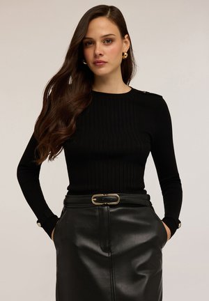 Femme aux longs cheveux bruns portant des boucles d'oreilles en or, un pull noir à côtes et une jupe en cuir noir avec une ceinture à boucle dorée, les mains dans les poches.