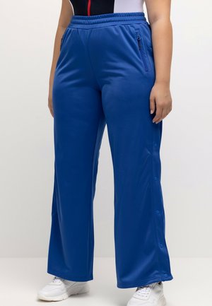 Persoon in een blauwe wijde sportbroek met drukknoopjes aan de zijkanten, gecombineerd met witte sneakers en een mouwloos shirt.
