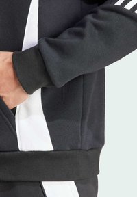 Persona che indossa un felpa grigia scura con strisce bianche sui lati, mano sinistra appoggiata nella tasca frontale.