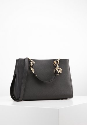 Sac à main en cuir noir avec des poignées en chaîne dorées et foncées, une longue bandoulière et un pendentif rond doré avec logo sur une surface blanche.