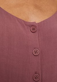 Camisa de lino burdeos con escote en V y cuatro botones a juego. La tela tiene una textura sutil con una apariencia suave y ligera.