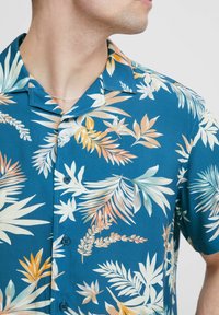 Chemise à manches courtes, boutonnée, en turquoise avec un motif de feuilles tropicales en blanc, orange et vert. Dotée d'un col standard et d'une texture lisse.