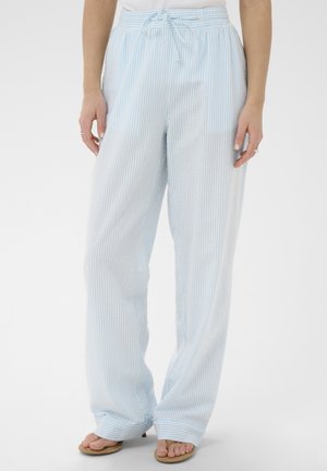 Femme portant un pantalon large à rayures bleu clair et blanc avec une taille élastique et un cordon de serrage, associé à des tongs, sur un fond uni.