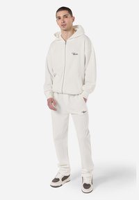 White ribbed hoodie with full zipper, side pockets, and logo. Matched with beige ribbed pants, featuring an elastic waistband and logo detail.