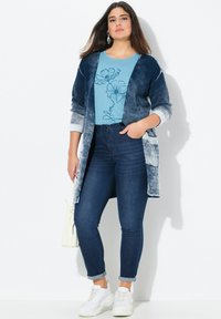 Blauer Verlauf Cardigan über einem blumigen blauen T-Shirt, kombiniert mit dunklen Skinny Jeans und weißen Sneakers. Inklusive einer cremefarbenen Handtasche.