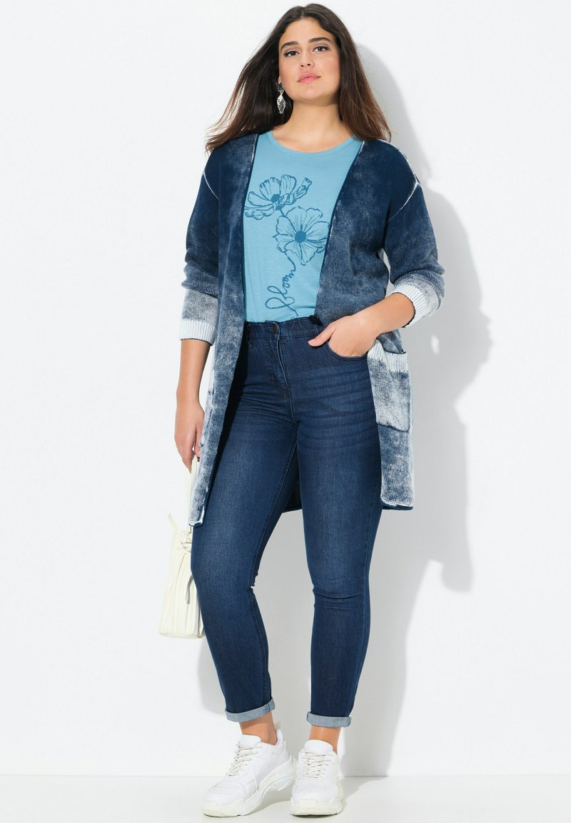 Blauer Verlauf Cardigan über einem blumigen blauen T-Shirt, kombiniert mit dunklen Skinny Jeans und weißen Sneakers. Inklusive einer cremefarbenen Handtasche.