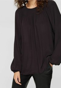 Blouse noire fluide à manches longues, au design plissé et aux poignets élastiqués, associée à un pantalon noir ajusté. Texture de tissu lisse.