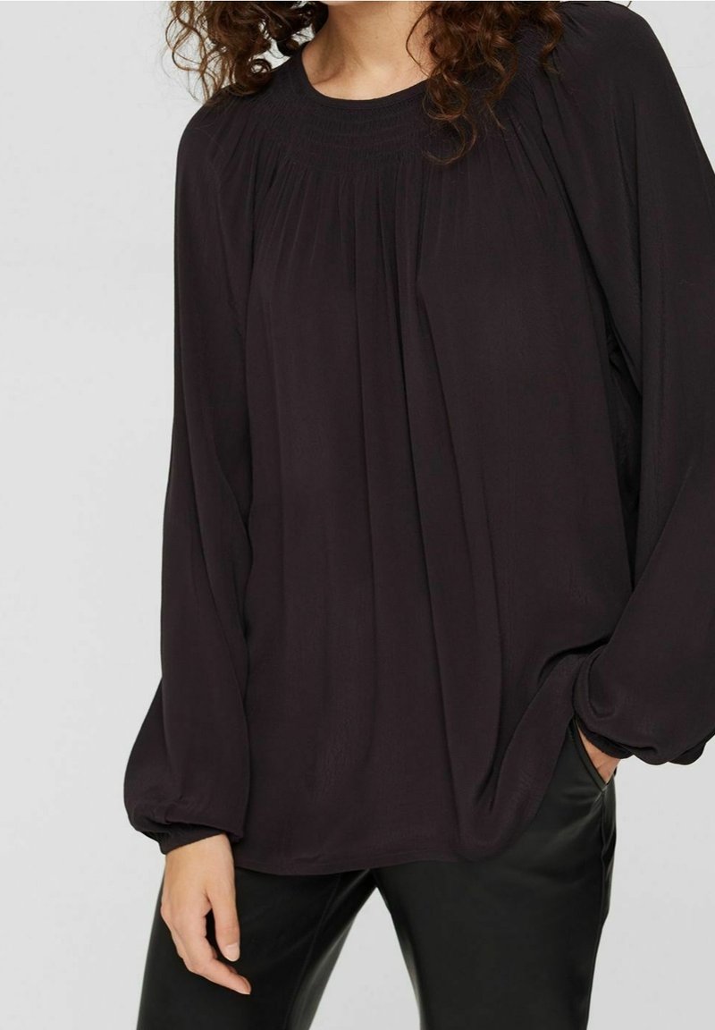 Blouse noire fluide à manches longues, au design plissé et aux poignets élastiqués, associée à un pantalon noir ajusté. Texture de tissu lisse.