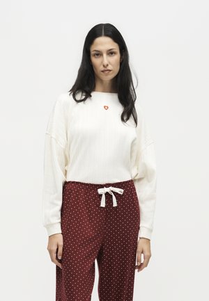 Kvinna med långt mörkt hår som bär en vit texturerad tröja och vinröda prickiga byxor med ett vitt dragsko rosett.