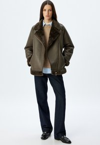 Brązowa kurtka z owczej wełny z teksturowanej skóry, z szerokim kołnierzem i zamkiem błyskawicznym, noszona na beżowym swetrze i jasnoniebieskiej koszuli, w zestawieniu z ciemnymi dżinsami.