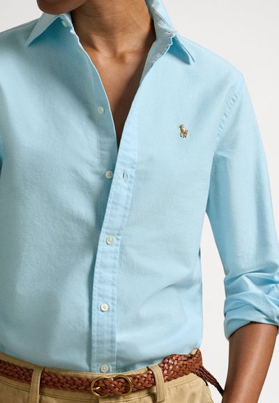 Gaiši zila krekla ar klasisku apkakli, vieglu audumu un izšūtu logotipu uz krūtīm. Piedurknes uzlocītas, nēsātas ar austu jostu.