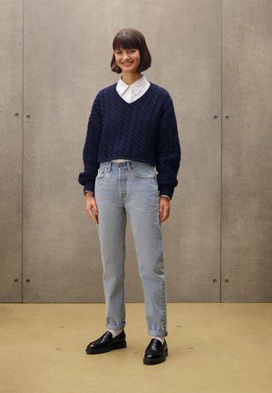 Suéter recortado de punto color navy con escote en V, combinado con jeans de tiro alto en azul claro y mocasines negros, de pie frente a una pared texturizada.
