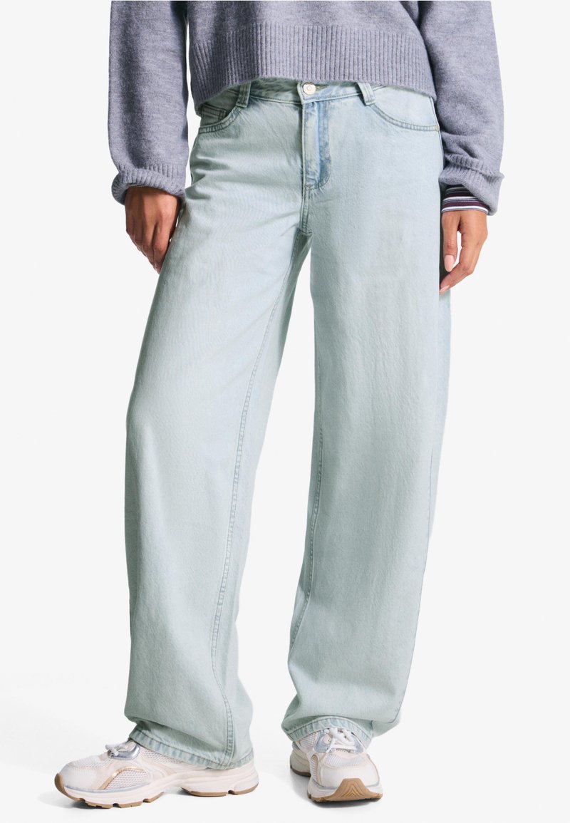 Lichtblauwe wijde spijkerbroek van denim, met een knoopsluiting en voorzakken. Gepaard met witte sneakers. Eenvoudig, casual ontwerp.