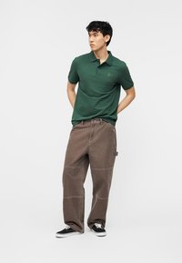 Νέος άνδρας στέκεται με τα χέρια πίσω από την πλάτη, φοράει πράσινο πόλο μπλουζάκι, χαλαρό καφέ παντελόνι και μαύρα αθλητικά παπούτσια, μπροστά σε απλό λευκό φόντο.