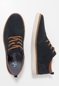 Scarpe in suede navy con lacci e dettagli marroni. Presentano una suola in gomma bianca con scanalature e cuciture lungo il bordo.