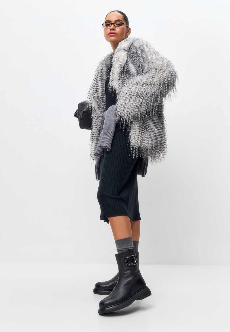 Abrigo gris y difuso con flecos largos, vestido negro de canalé, guantes de punto gris y botines negros con detalle de hebilla. Accesorios simples y modernos.