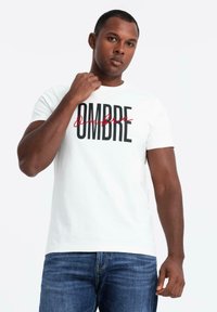 Camiseta de algodón blanca con texto en negrita negro "OMBRE" y un acento en cursiva rojo. Manga corta, cuello redondo y ajuste relajado.