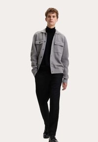 Szary kardigan o ryflowanej strukturze, z dwiema kieszeniami na piersi i czarnymi guzikami, noszony na czarnym golfie i czarnych spodniach, z eleganckimi czarnymi butami.