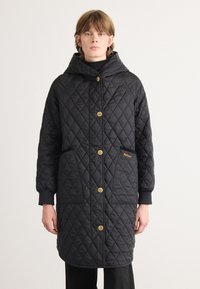 Zwarte gewatteerde lange jas met capuchon, voorzien van gouden knoopsluitingen, zijzakken en een subtiele logo op de zak. Glad textuur.