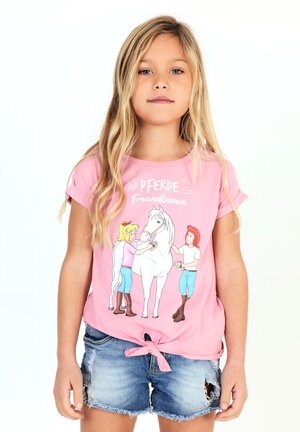 Junges Mädchen mit langen blonden Haaren trägt ein rosa geknotetes Shirt mit zwei Mädchen, die ein weißes Pferd pflegen, und Jeansshorts.