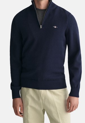 Mężczyzna w granatowym swetrze z krótkim zamkiem, z prążkowanym kołnierzem, mankietami i pasem, w połączeniu z jasnobeżowymi spodniami, na jednolitym tle.